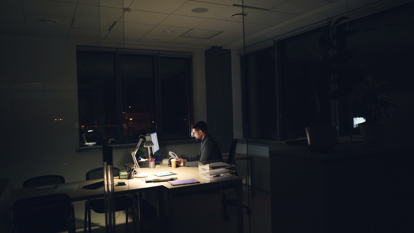 A solo builder working at a desk lamp in a dim office at night, wide shot